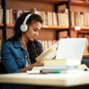 A woman sitting at a computer reads a book with headphones on in a library.