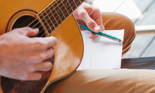 A musician holds a guitar in their right hand and notes with a pen in their left hand.