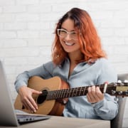 A young woman plays the guitar while interacting with another person via her computer.