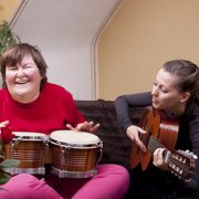 A woman with special needs plays the drums and laughs while another woman sings and plays the guitar.