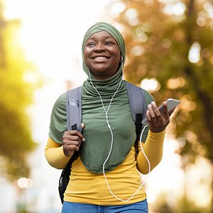 Happy young woman in a hijab listening to music with smartphone and headphones while walking outdoors with a backpack.